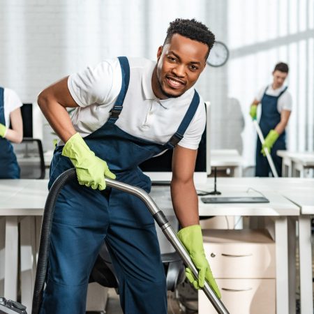 cheerful african american cleaner vacuuming floor while looking at camera